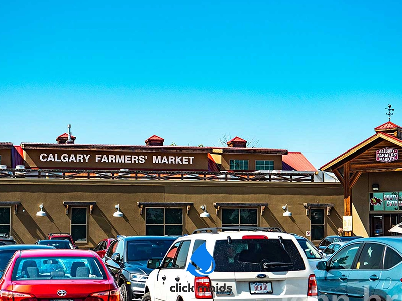 "Calgary Farmers' Market exterior with brown facade, red roof, bold signage, parked cars, and clear blue sky."
