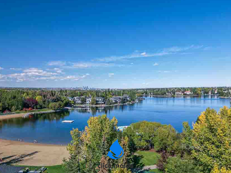 Aerial view of McKenzie Lake, Calgary, with beach area, homes, and the city skyline under a blue sky.
