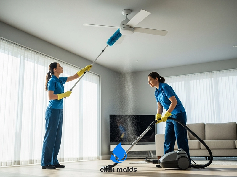 "Two cleaners in blue uniforms and yellow gloves dusting ceiling fan and vacuuming floor in sunlit living room with sheer curtains and white sofa, 'click maids' logo below."