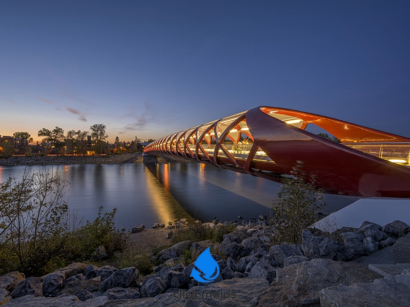 Peace Bridge in Calgary glows red at twilight over Bow River; glass panels reflect blue sky and riverbank rocks below.