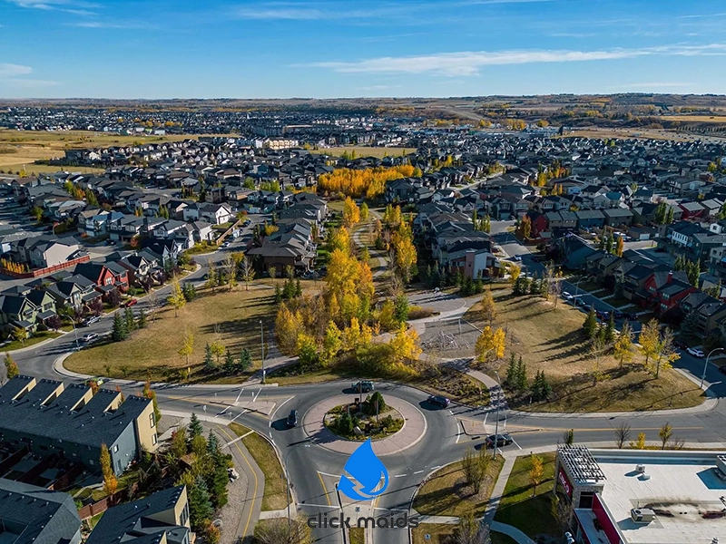 "Aerial view of suburban roundabout with autumn trees, surrounded by neat rows of homes and open fields, 'click maids' logo below."