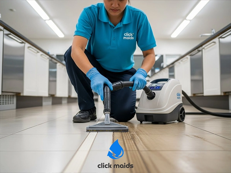 Professional cleaner wearing gloves uses a floor-cleaning vacuum to deep clean tiled flooring in a commercial kitchen or workspace.