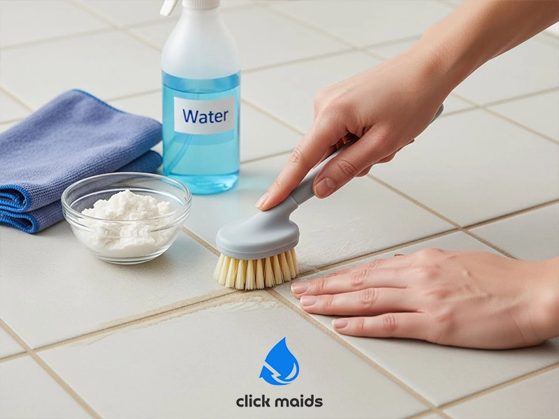 Hand scrubbing tile grout with a small brush, using water and cleaning powder, with a spray bottle and cloth nearby.