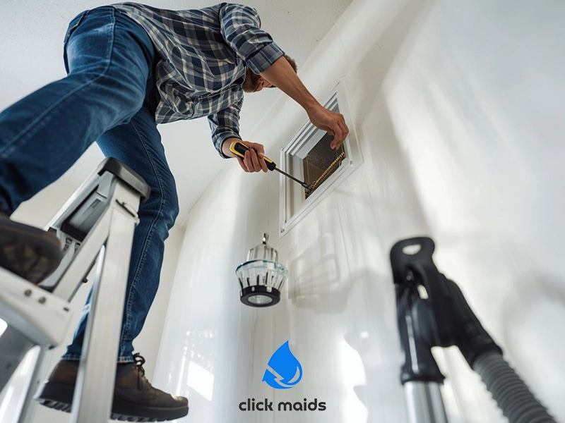 Man standing on a step ladder while removing or cleaning a wall air vent using a screwdriver. A vacuum cleaner hose is positioned nearby, ready to collect dust and debris. The bright room with white walls suggests routine home maintenance focused on improving indoor air quality.