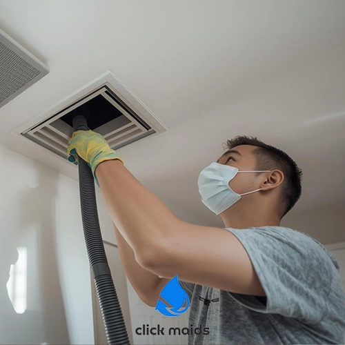 Click Maids Professional cleaner wearing a mask and gloves vacuuming a ceiling air vent as part of residential duct cleaning.