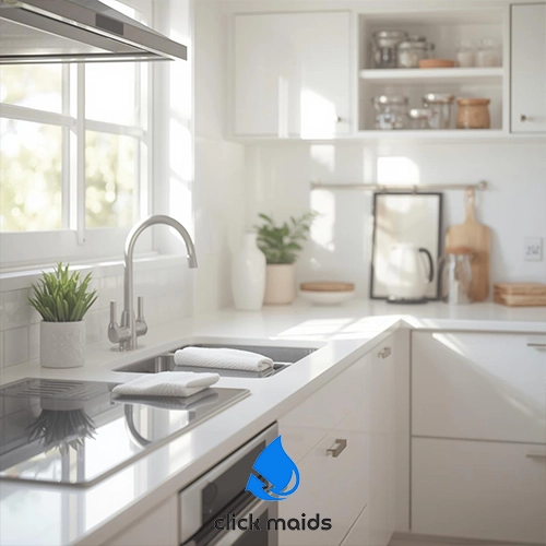 Click Maids Bright, modern white kitchen with sunlight streaming through a large window above the sink, featuring clean countertops, folded towels, open shelving with neatly arranged jars, potted plants, and a minimalist, clutter-free design.